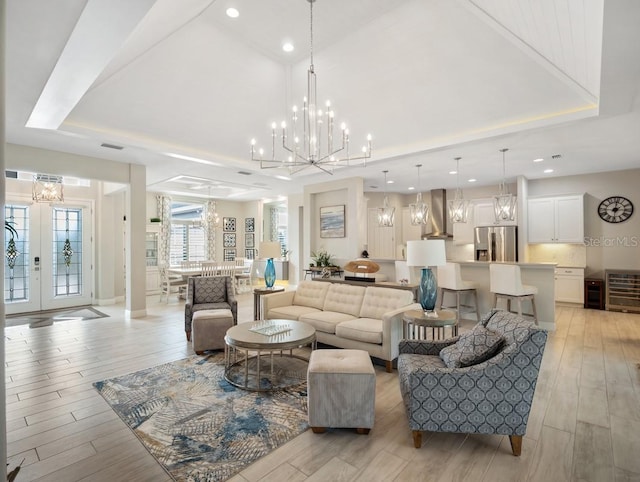 living room featuring a notable chandelier, beverage cooler, light wood-style floors, french doors, and a tray ceiling