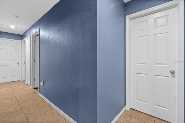 hall with a textured wall, a textured ceiling, baseboards, and light tile patterned floors