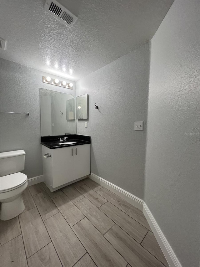 bathroom with a textured wall, toilet, vanity, visible vents, and baseboards