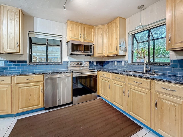 kitchen featuring stainless steel appliances, a sink, backsplash, dark stone counters, and light brown cabinetry