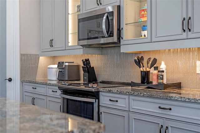 kitchen featuring stainless steel appliances, glass insert cabinets, light stone counters, and decorative backsplash