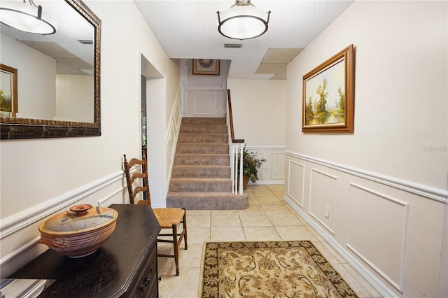 corridor featuring visible vents, a textured ceiling, a decorative wall, light tile patterned floors, and stairs