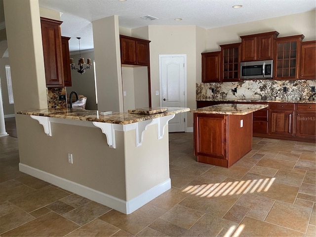 kitchen featuring tasteful backsplash, stainless steel microwave, a peninsula, stone tile flooring, and a kitchen bar