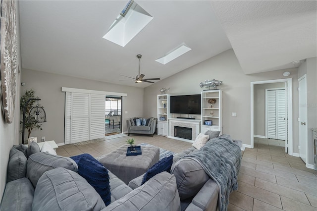 living area featuring a fireplace, a ceiling fan, light tile patterned flooring, vaulted ceiling with skylight, and baseboards