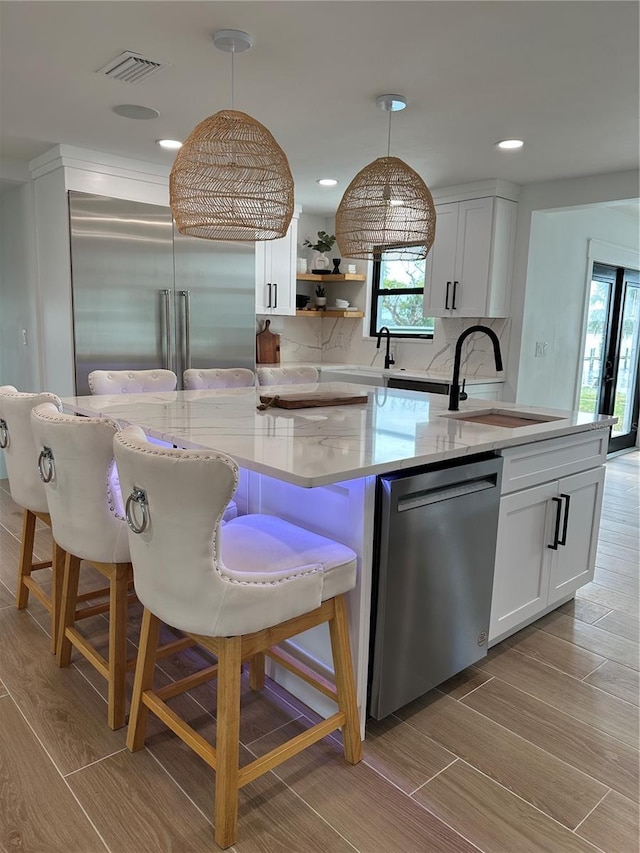 kitchen with a center island with sink, wood finish floors, a sink, stainless steel appliances, and pendant lighting