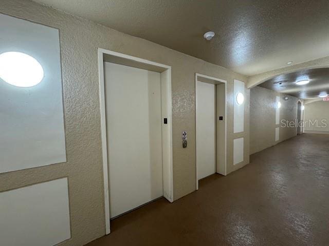 hall with concrete floors, elevator, arched walkways, a textured ceiling, and a textured wall