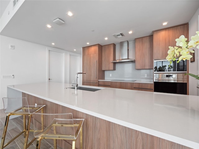 kitchen with brown cabinetry, a sink, stainless steel oven, wall chimney range hood, and black electric cooktop