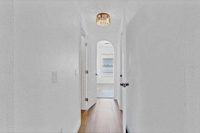 hallway with arched walkways, a textured wall, baseboards, and light wood-style floors