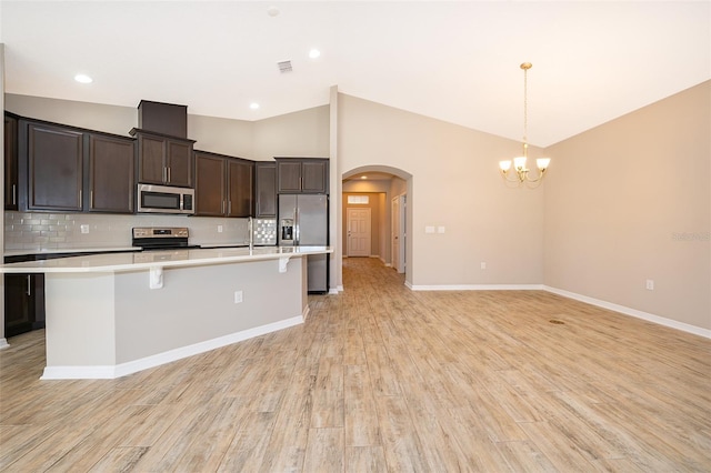 kitchen featuring arched walkways, a center island with sink, stainless steel appliances, light countertops, and dark brown cabinetry