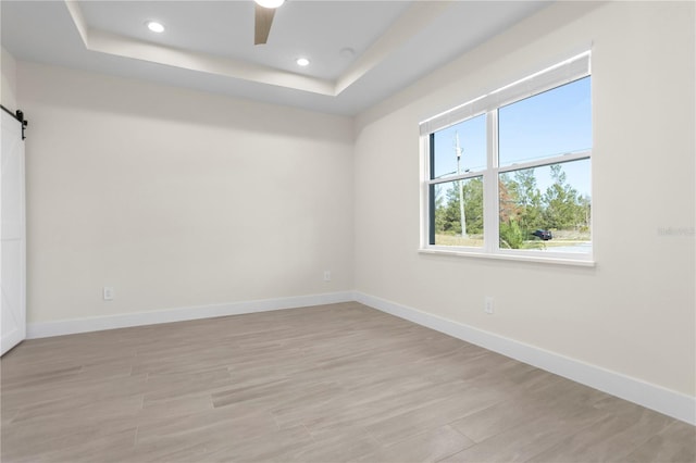 empty room featuring a barn door, baseboards, a tray ceiling, light wood-style floors, and recessed lighting