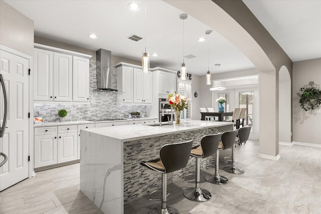 kitchen with arched walkways, black electric stovetop, a kitchen island with sink, a breakfast bar, and wall chimney exhaust hood