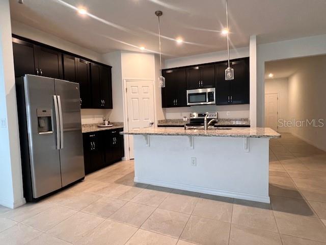 kitchen with light stone counters, light tile patterned flooring, hanging light fixtures, appliances with stainless steel finishes, and a kitchen bar