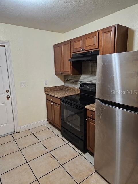 kitchen with light tile patterned floors, black electric range oven, freestanding refrigerator, a textured ceiling, and under cabinet range hood