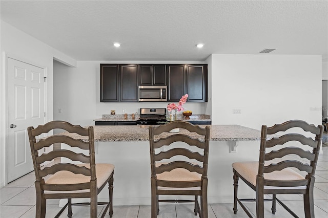 kitchen with a textured ceiling, appliances with stainless steel finishes, a kitchen bar, and light stone countertops