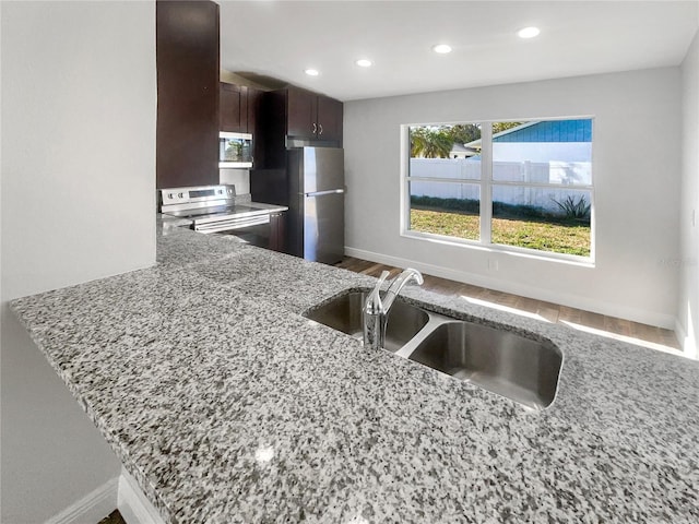 kitchen with dark brown cabinetry, light stone counters, stainless steel appliances, a sink, and recessed lighting