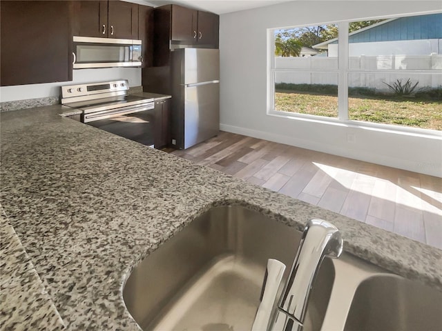 kitchen featuring baseboards, appliances with stainless steel finishes, dark brown cabinets, light wood-type flooring, and stone counters