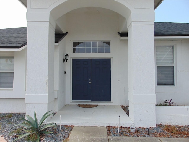 view of exterior entry with stucco siding and a shingled roof