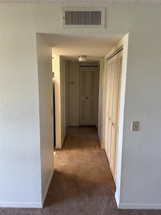 hallway with a textured wall, carpet, visible vents, and baseboards