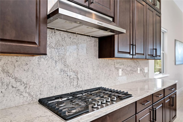 kitchen featuring stainless steel gas cooktop, backsplash, dark brown cabinetry, light stone countertops, and under cabinet range hood