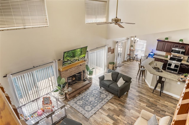 living room featuring light wood finished floors, a brick fireplace, baseboards, ceiling fan with notable chandelier, and a towering ceiling