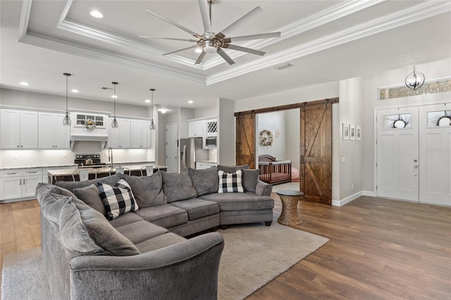 living room featuring a barn door, visible vents, a raised ceiling, ornamental molding, and wood finished floors