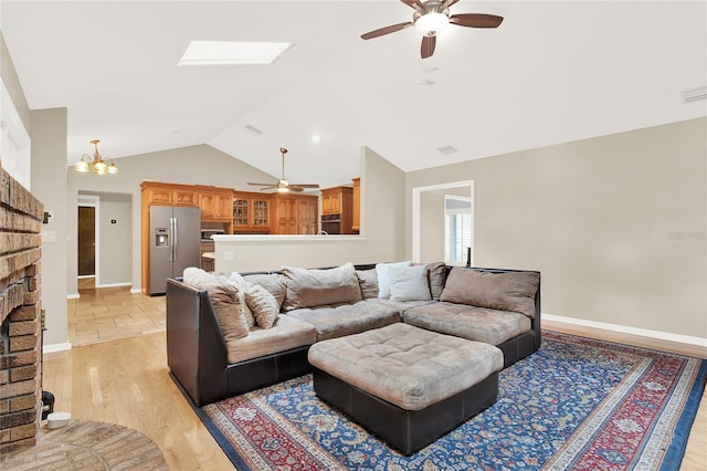 living area with baseboards, vaulted ceiling with skylight, light wood-style flooring, a fireplace, and ceiling fan with notable chandelier