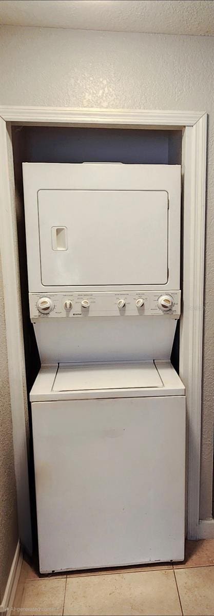 clothes washing area featuring a textured wall, stacked washer and dryer, light tile patterned flooring, and laundry area