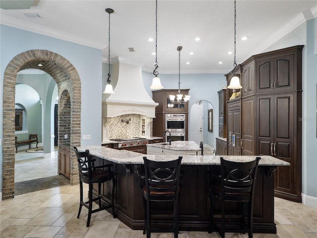 kitchen with arched walkways, custom exhaust hood, decorative backsplash, ornamental molding, and a large island with sink