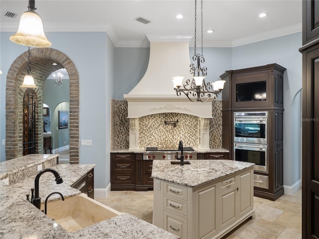 kitchen featuring arched walkways, double oven, a kitchen island with sink, a sink, and visible vents