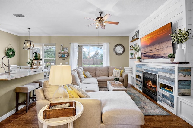 living area featuring ceiling fan, a stone fireplace, dark wood-type flooring, visible vents, and crown molding