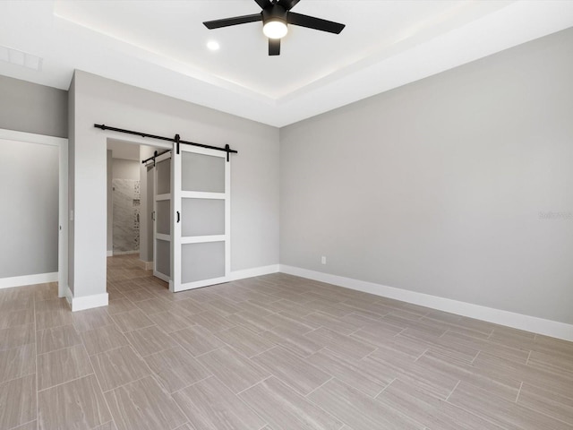 unfurnished bedroom with a tray ceiling, a barn door, wood tiled floor, ceiling fan, and baseboards