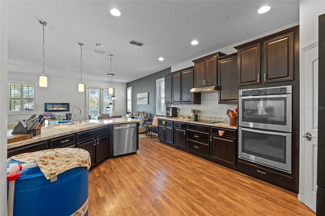 kitchen with under cabinet range hood, light wood-type flooring, light stone counters, appliances with stainless steel finishes, and a sink