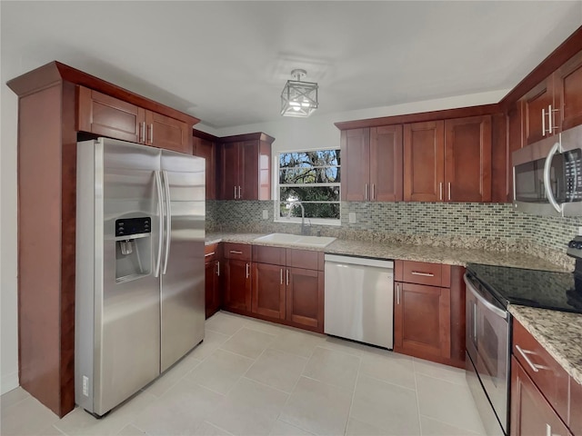 kitchen with stainless steel appliances, a sink, decorative backsplash, light stone countertops, and reddish brown cabinets