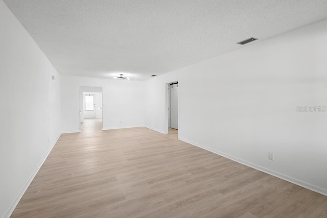 empty room with light wood finished floors, visible vents, a barn door, a textured ceiling, and baseboards