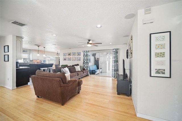 living room featuring a textured ceiling, visible vents, baseboards, a ceiling fan, and light wood finished floors