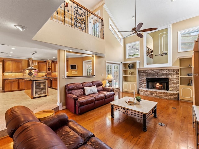 living room with light wood finished floors, a ceiling fan, wine cooler, ornamental molding, and a brick fireplace