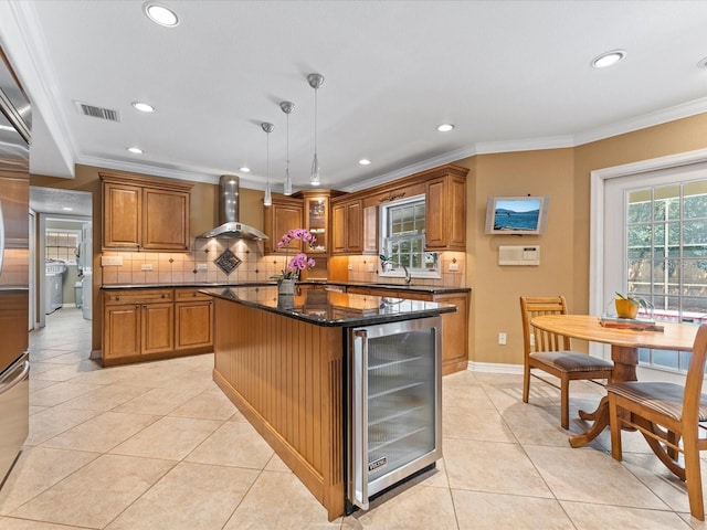 kitchen with light tile patterned floors, wine cooler, brown cabinets, crown molding, and wall chimney range hood