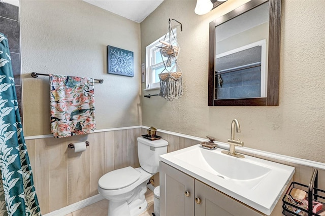 full bathroom with a wainscoted wall, a textured wall, a shower with shower curtain, toilet, and vanity