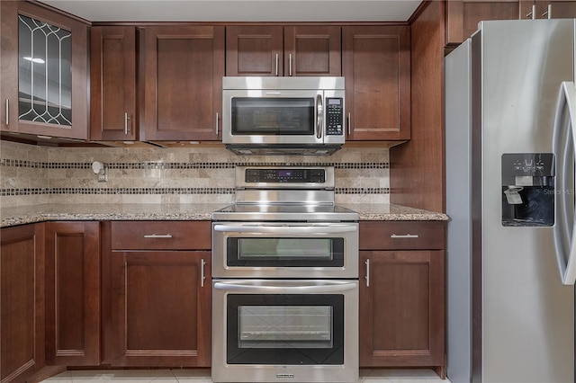 kitchen featuring stainless steel appliances, glass insert cabinets, light stone countertops, and decorative backsplash