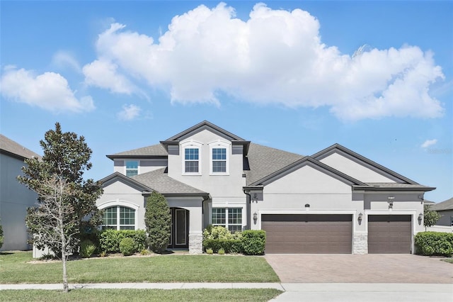 traditional home with an attached garage, a shingled roof, stucco siding, a front lawn, and decorative driveway