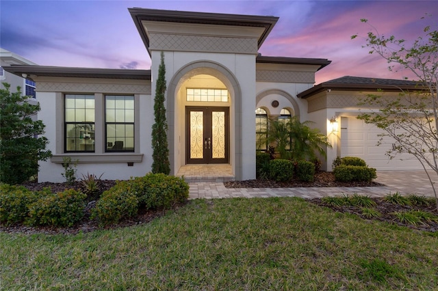 exterior entry at dusk with french doors, stucco siding, a lawn, an attached garage, and driveway