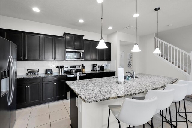 kitchen featuring light stone counters, a center island with sink, tasteful backsplash, appliances with stainless steel finishes, and dark cabinets