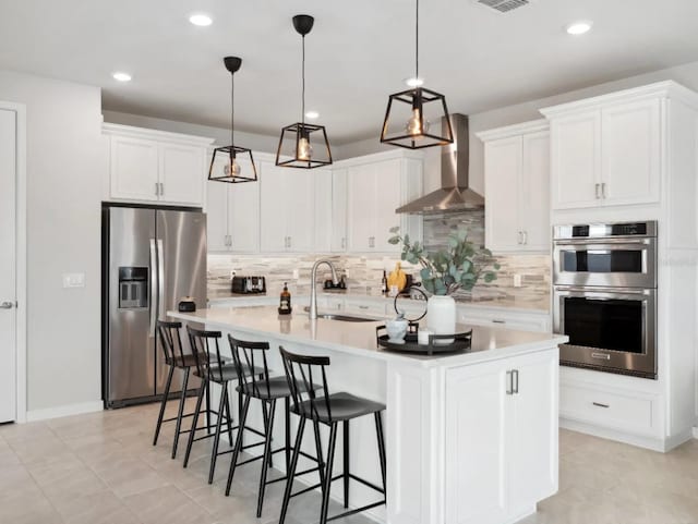 kitchen with a center island with sink, stainless steel appliances, tasteful backsplash, a sink, and wall chimney exhaust hood
