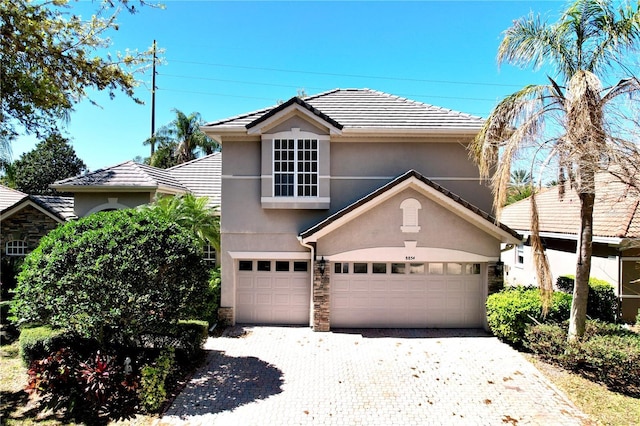 traditional-style house with stucco siding, a garage, stone siding, a tiled roof, and decorative driveway