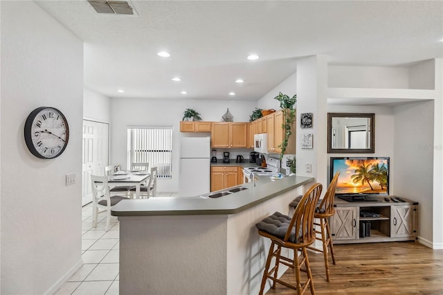 kitchen featuring visible vents, light brown cabinetry, a breakfast bar, a peninsula, and white appliances