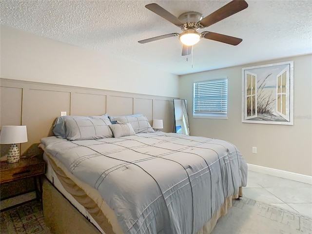 bedroom featuring a textured ceiling, ceiling fan, light tile patterned flooring, a decorative wall, and wainscoting