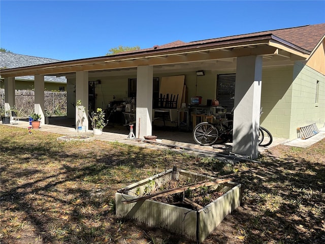 back of house featuring a shingled roof, concrete block siding, a patio area, and a vegetable garden