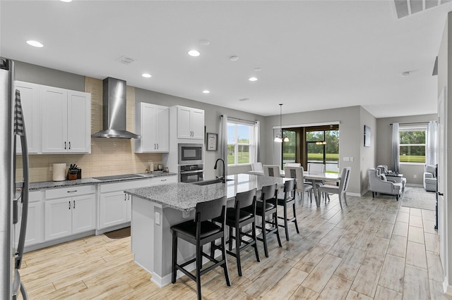 kitchen with a center island with sink, light wood-style flooring, wall chimney range hood, black appliances, and backsplash