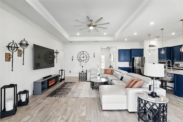 living area featuring light wood finished floors, a tray ceiling, a glass covered fireplace, recessed lighting, and ceiling fan
