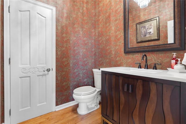 half bathroom featuring vanity, wood finished floors, baseboards, wallpapered walls, and toilet